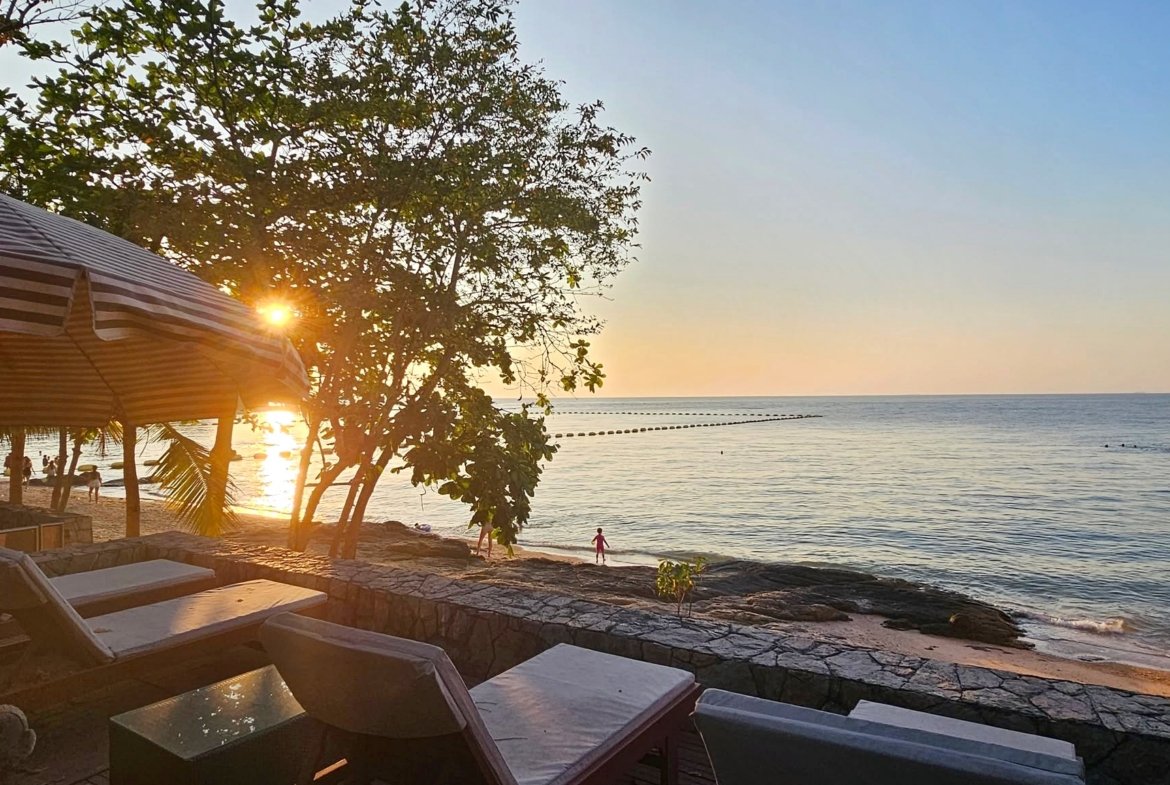 Sunset over the calm ocean with a striped canopy and lounge chairs in the foreground on a sandy beach. The sun shines through trees near the shore.