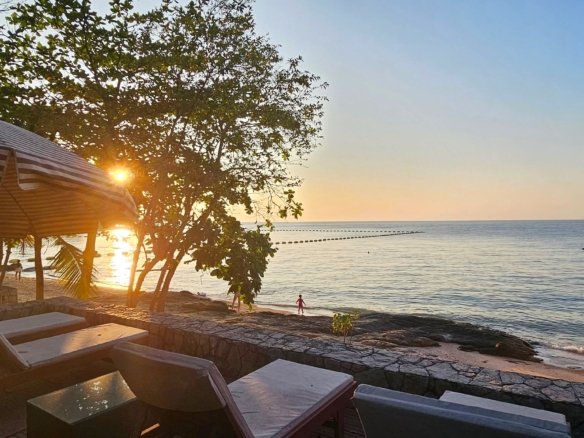 Sunset over the calm ocean with a striped canopy and lounge chairs in the foreground on a sandy beach The sun shines through trees near the shore
