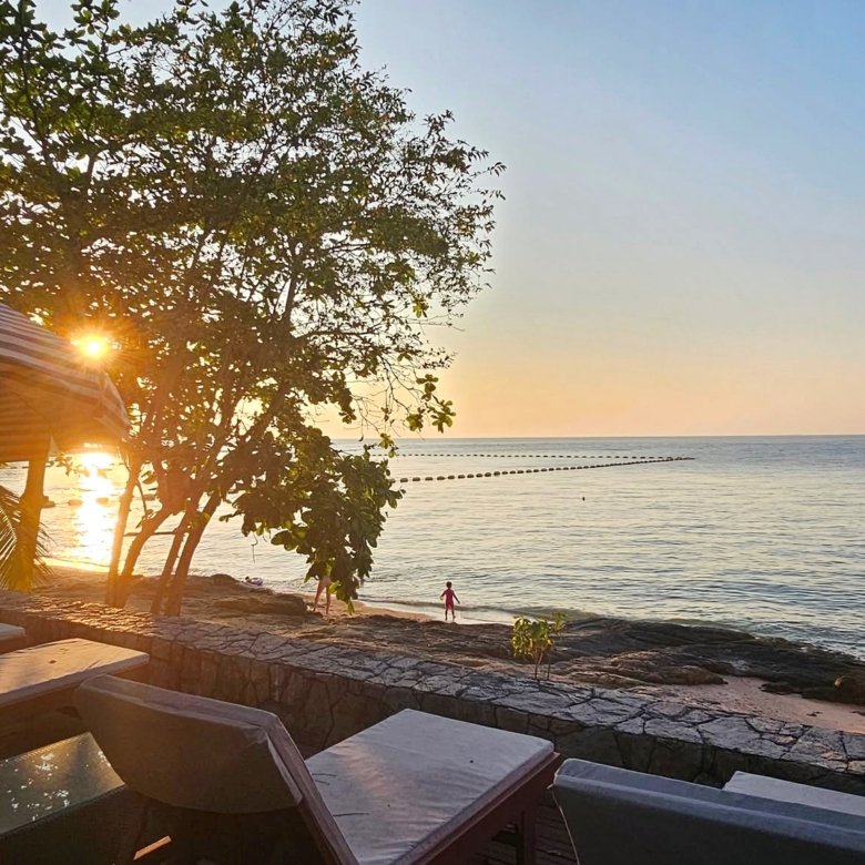 Sunset over the calm ocean with a striped canopy and lounge chairs in the foreground on a sandy beach The sun shines through trees near the shore