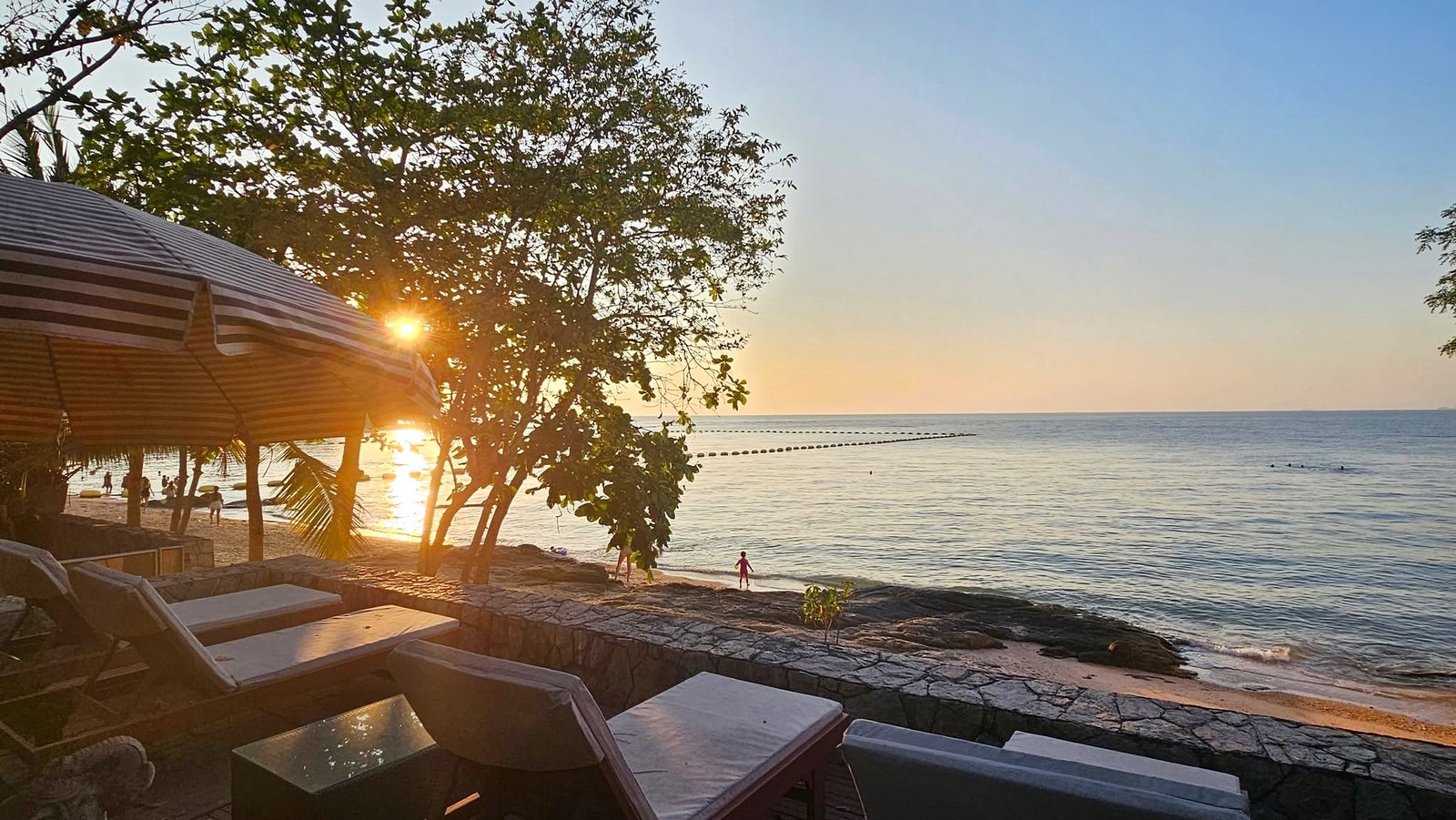 Wongamat_House_Rent_Rdl357_2 Sunset over the calm ocean with a striped canopy and lounge chairs in the foreground on a sandy beach. The sun shines through trees near the shore.