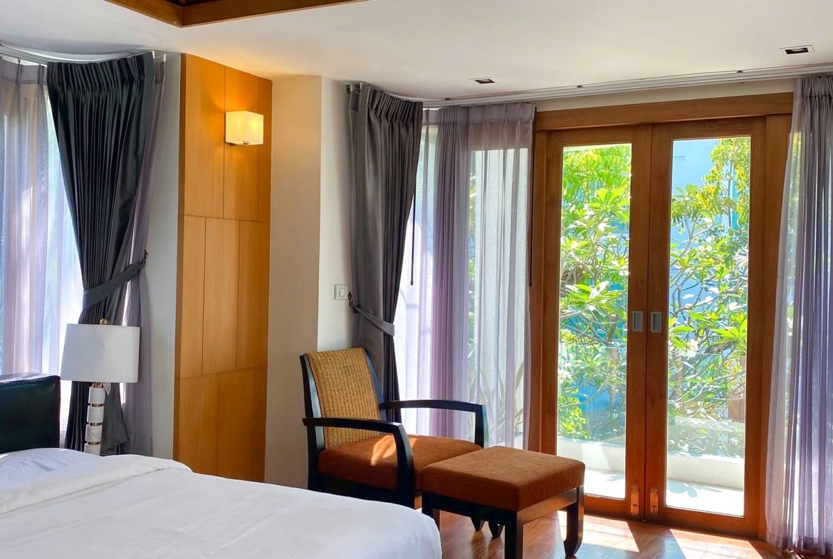 Hotel room with a white bed in the foreground, wooden ceiling beams, and glass doors opening to greenery outside.