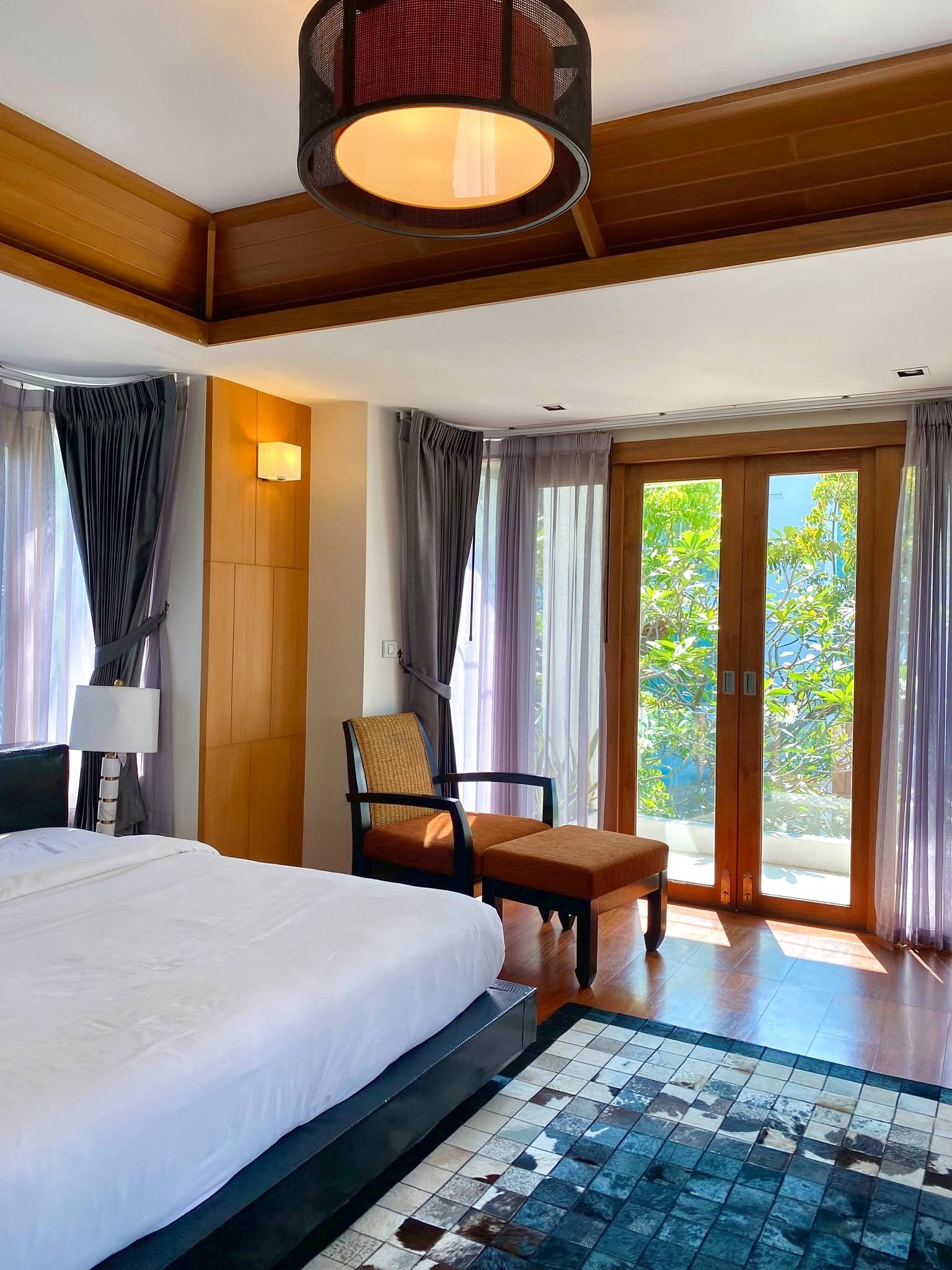 Wongamat_House_Rent_Rdl357_4 Hotel room with a white bed in the foreground, wooden ceiling beams, and glass doors opening to greenery outside.