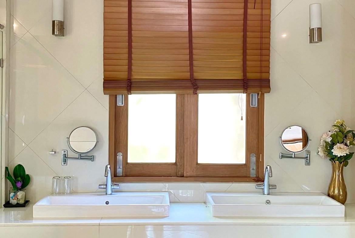 Double-sink bathroom vanity with white counters, chrome taps, and warm wooden cabinets beneath a window with brown blinds.
