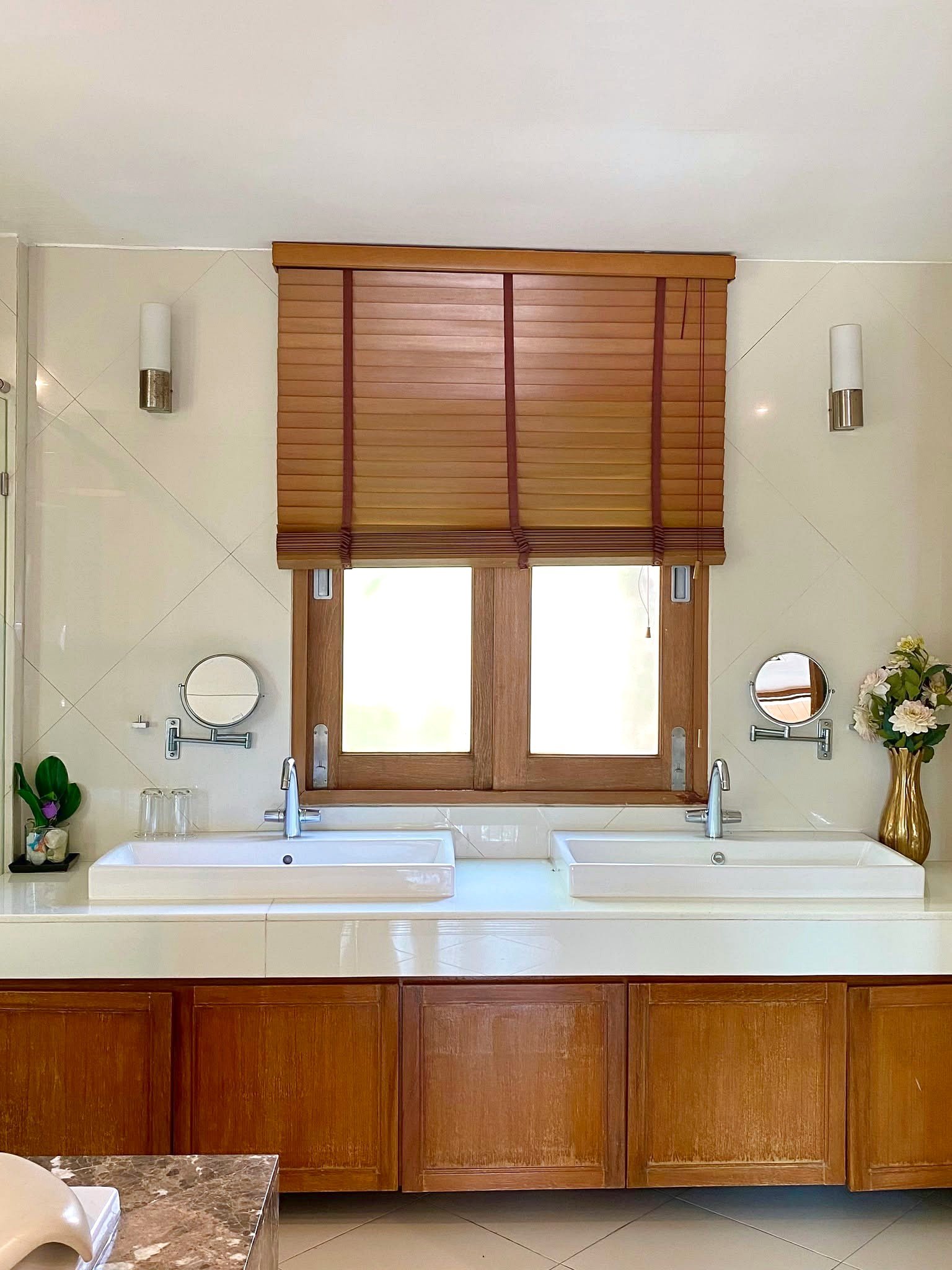 Wongamat_House_Rent_Rdl357_8 Double-sink bathroom vanity with white counters, chrome taps, and warm wooden cabinets beneath a window with brown blinds.