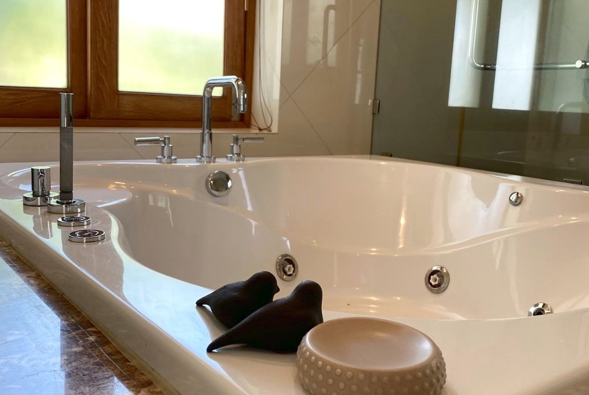 Luxurious white jacuzzi tub with chrome fixtures in a marble-floored bathroom, wood-framed window and glass shower in the background, a beige textured stool nearby.