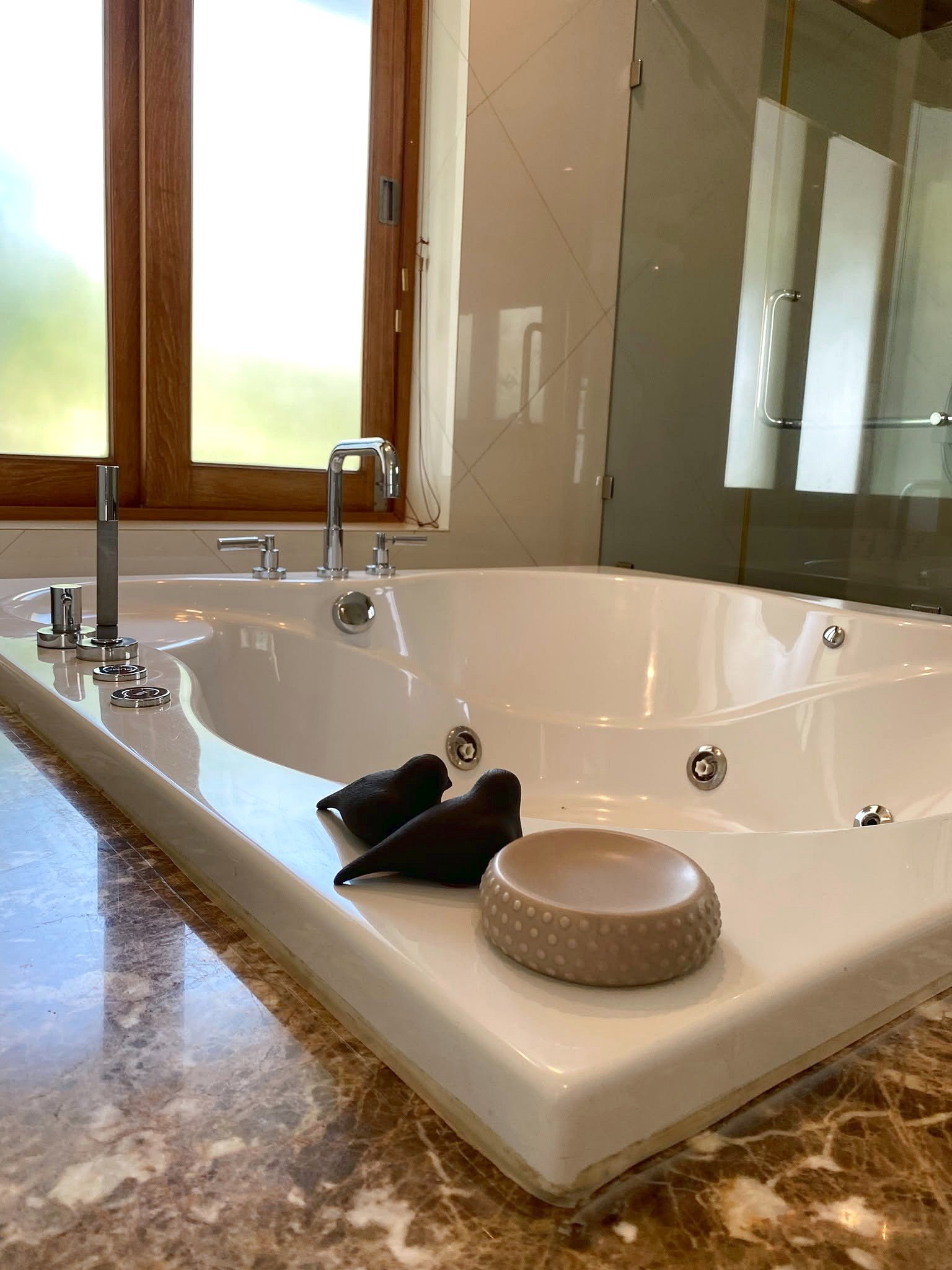 Wongamat_House_Rent_Rdl357_9 Luxurious white jacuzzi tub with chrome fixtures in a marble-floored bathroom, wood-framed window and glass shower in the background, a beige textured stool nearby.