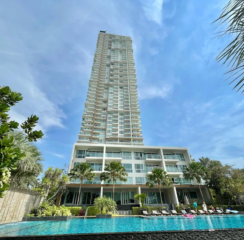 Tall white high rise hotel with balconies overlooking a resort style pool area and palm trees at the edge of a sunny day