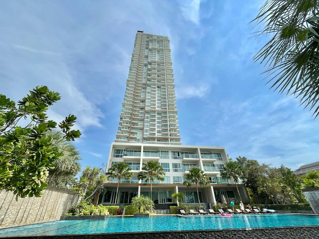 Tall white high-rise hotel with balconies, overlooking a resort-style pool area and palm trees at the edge of a sunny day.