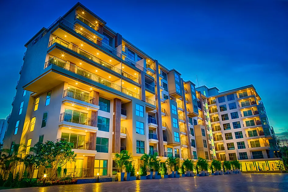 Modern apartment building at dusk, illuminated balconies and a landscaped foreground with palm trees.