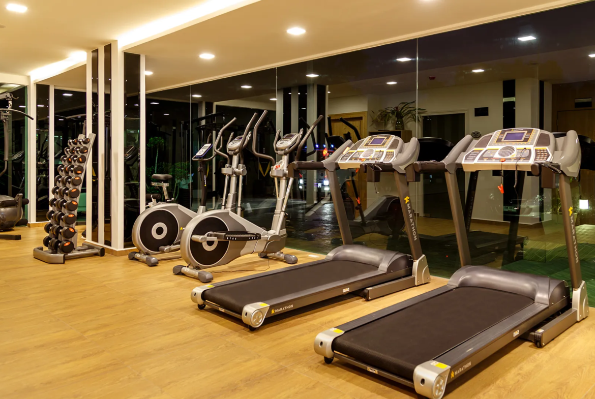 Well-lit gym with treadmills and elliptical machines along a mirrored wall and a rack of dumbbells nearby.