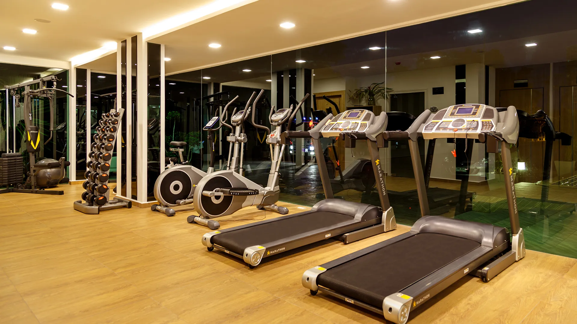 Well-lit gym with treadmills and elliptical machines along a mirrored wall and a rack of dumbbells nearby.