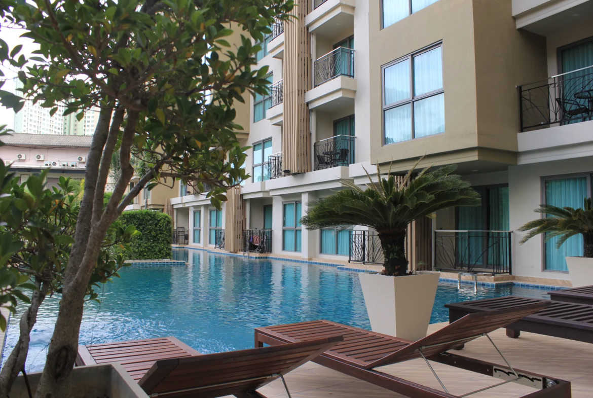 Hotel-style pool deck with a rectangular blue pool, lounge wooden chairs, and palm plants beside a beige apartment building with balconies and large windows.