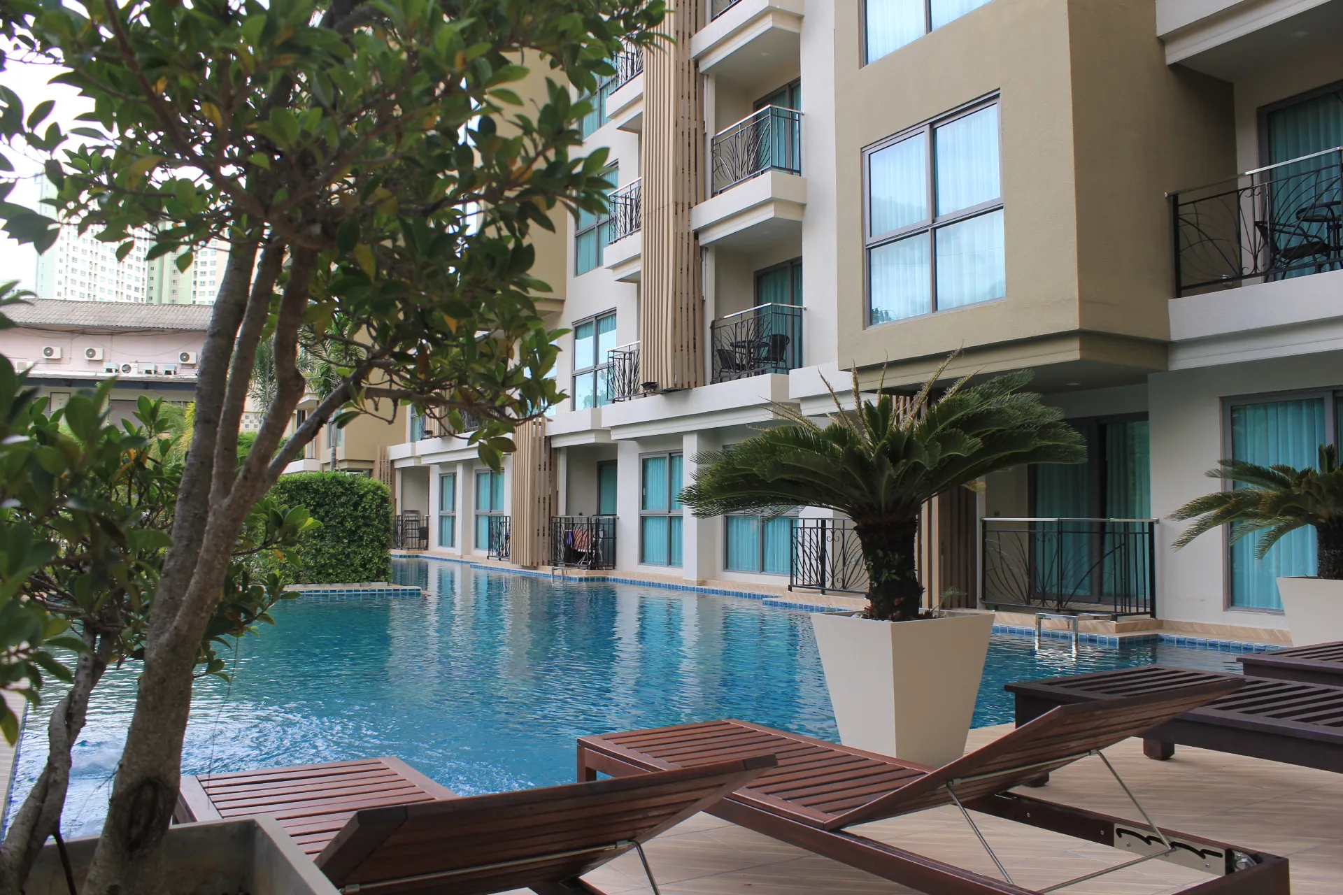 Hotel-style pool deck with a rectangular blue pool, lounge wooden chairs, and palm plants beside a beige apartment building with balconies and large windows.