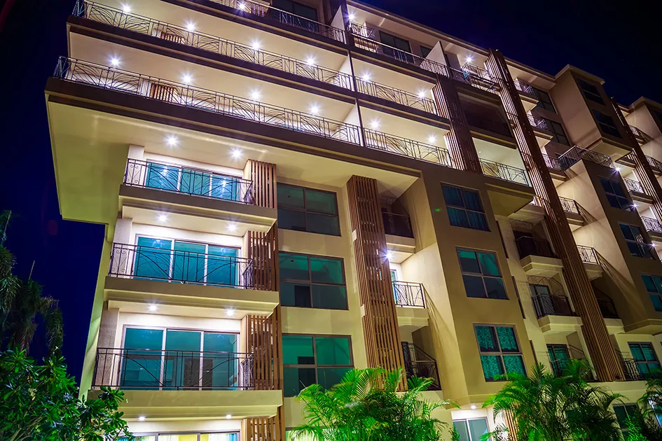 Night view of a modern multi-story apartment building with illuminated balconies and palm trees in the foreground.